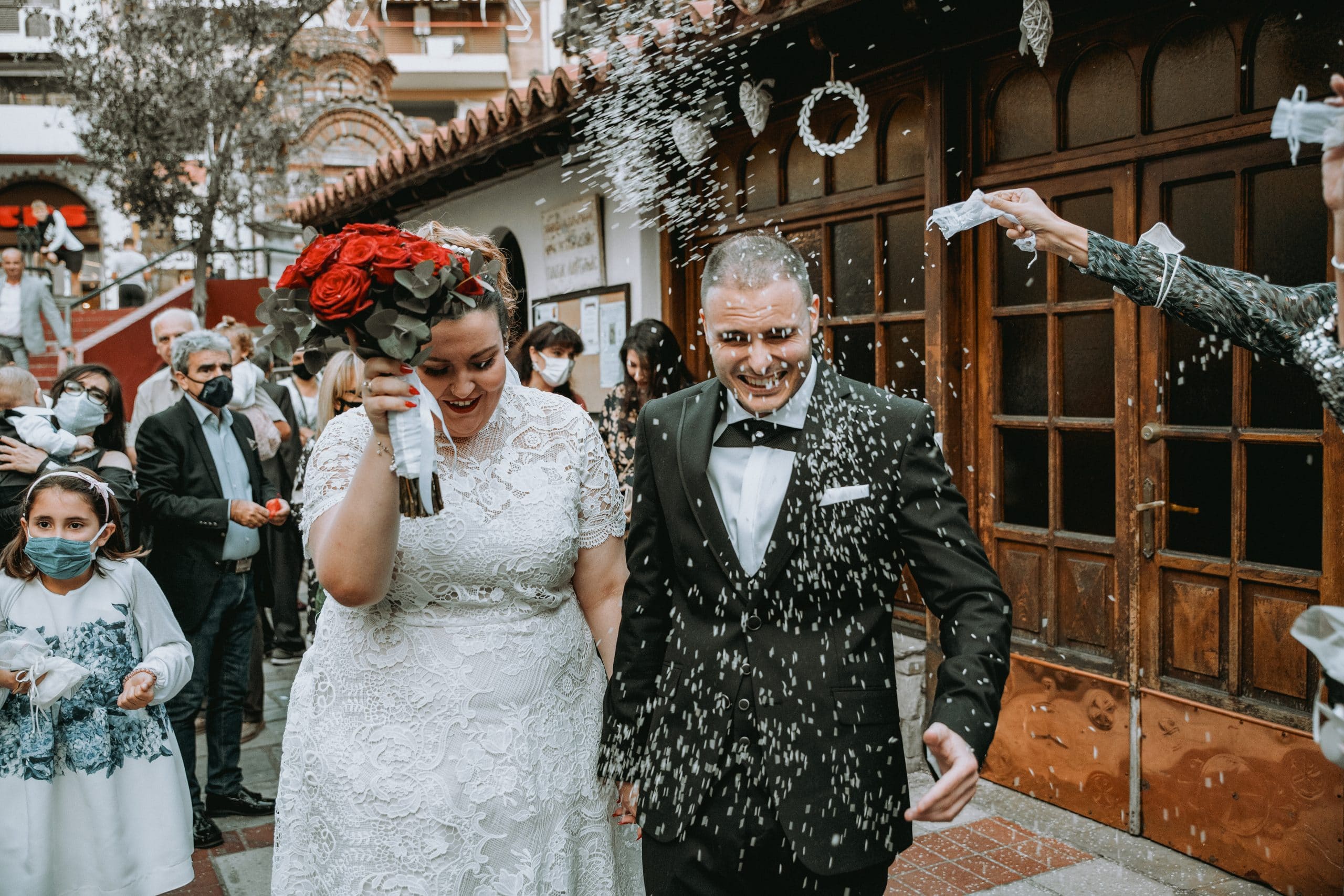 Φωτογραφία κατα την φωτογράφιση γάμου στην Θεσσαλονίκη, το ζευγάρι βγαίνει απο την εκκλησία και οι καλεσμένοι ρίχνουν ρύζι . Στην συγκεκριμένη φωτογραφία φαίνεται η χαρά της στιγμής . Ο γαμπρός φορά ενα μαυρο boss κοστούμι και η νύφη ενα ολόλευκο νυφικό και κρατά μια ανθοδέσμη με κόκκινα άνθη. Φωτογράφος Γιώργος Καρασαββίδης gkphotography